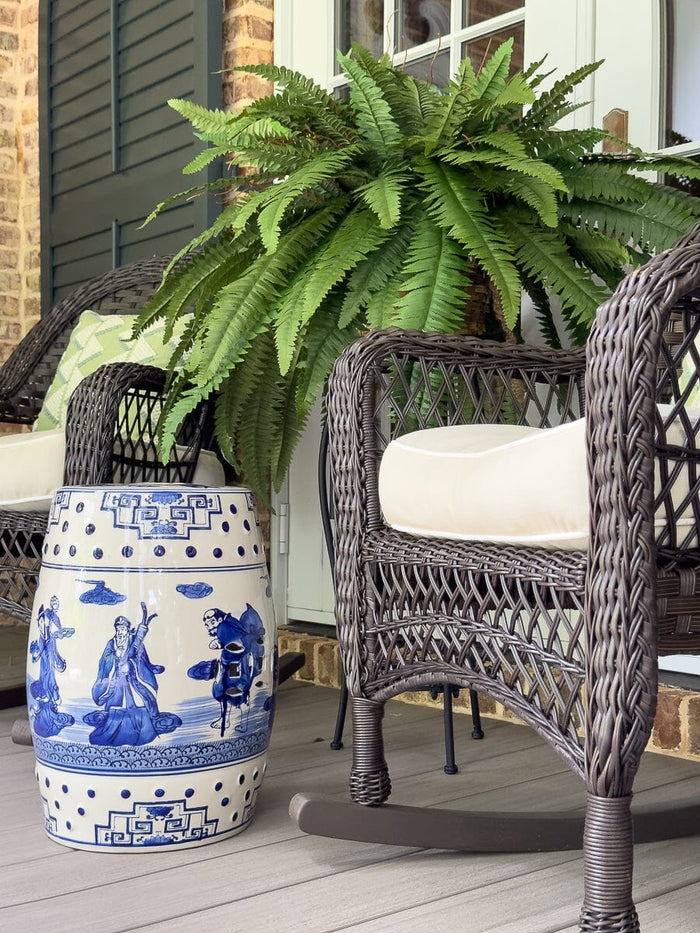 Wicker chair with a white cushion on a wooden deck, next to a large green fern and a decorative blue and white garden stool.